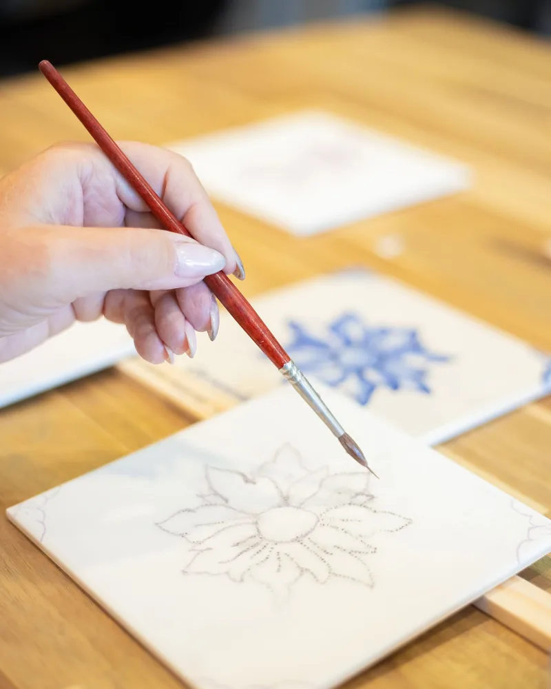 Mano pintando con pincel un motivo floral azul sobre un azulejo cerámico